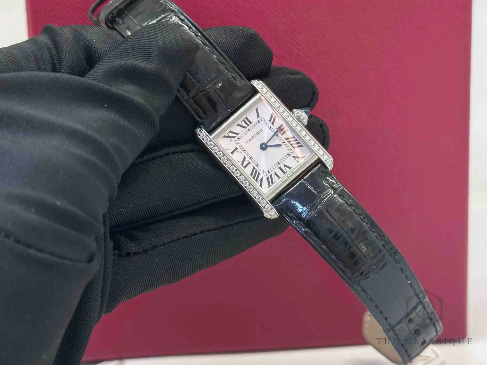 Đồng hồ Cartier cầm trên tay có găng, mặt số và kim xanh thép nhìn nghiêng dưới ánh đèn